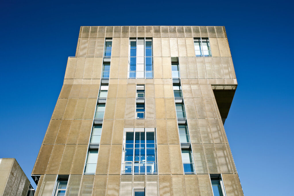 Beige gelbliche Fasssade aus transluzenten GFK-Gitterrosten. Die Fassade wird durch einige Fensterreihen unterbrochen und hat ein asymmetrisches ERscheinungsbild