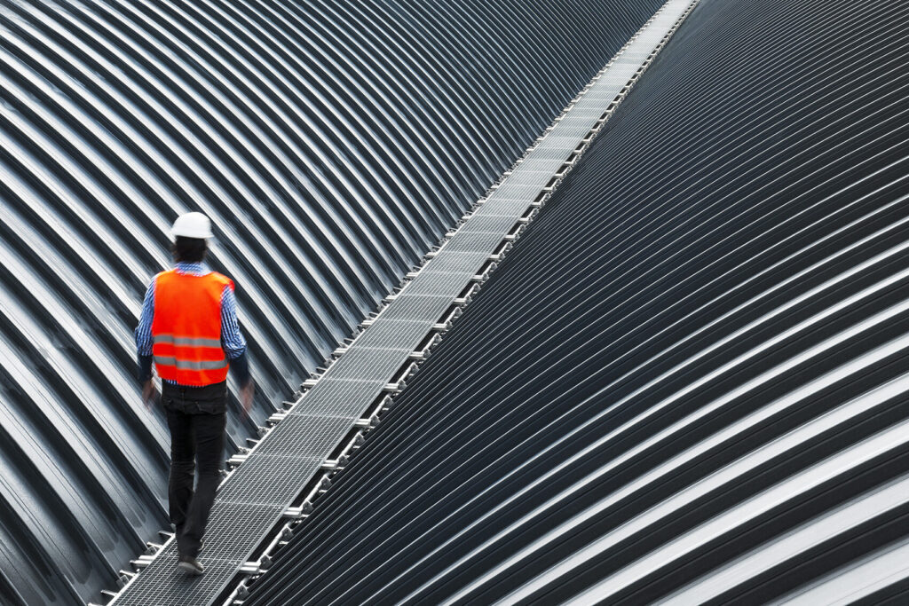 Wellblechdach eines Bahnhofes. Zwischen zwei Dächern befindet sich ein Laufsteg aus Gitterrosten.
